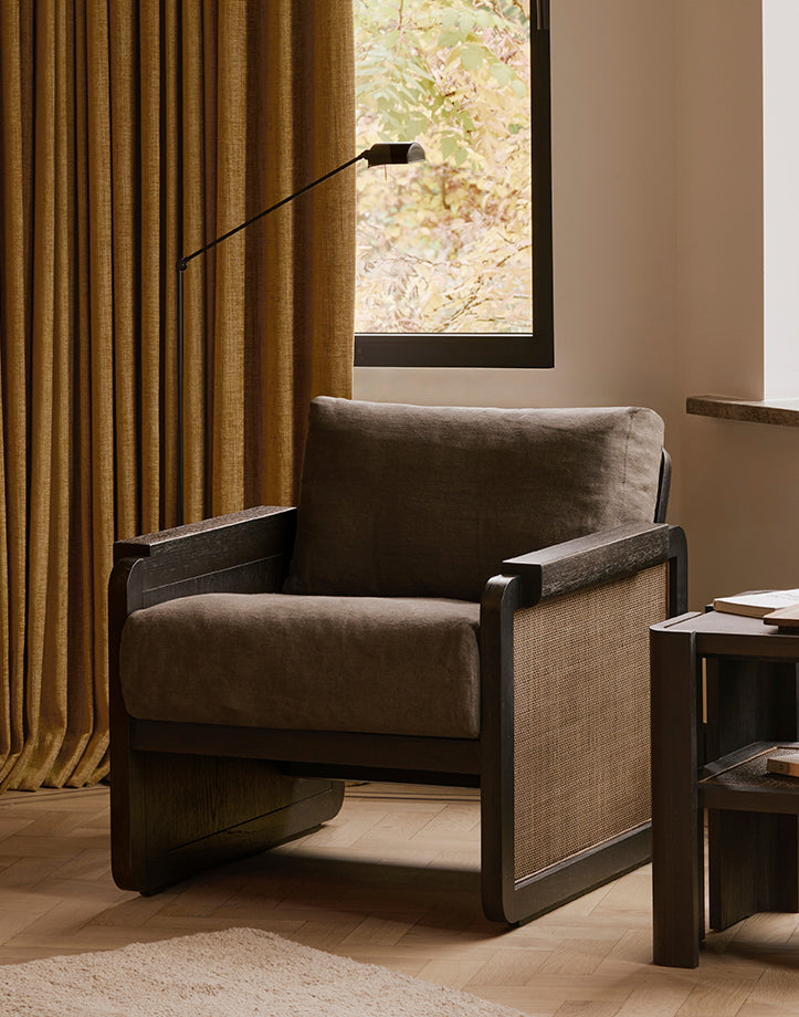 The Panel Lounge Chair - Dark Brown Teak by Ethnicraft, featuring a dark wood frame, brown cushion, and woven side panels, sits beside gold curtains and a side table in a cozy living room.