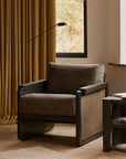 The Panel Lounge Chair - Dark Brown Teak by Ethnicraft, featuring a dark wood frame, brown cushion, and woven side panels, sits beside gold curtains and a side table in a cozy living room.