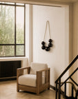 The Roan Lounge Chair | Flax by Ethnicraft, featuring solid teak construction and light-toned upholstery, is positioned next to a large window and radiator; above it hangs a wall sculpture adorned with black spheres, while a section of staircase with a black railing appears on the right.