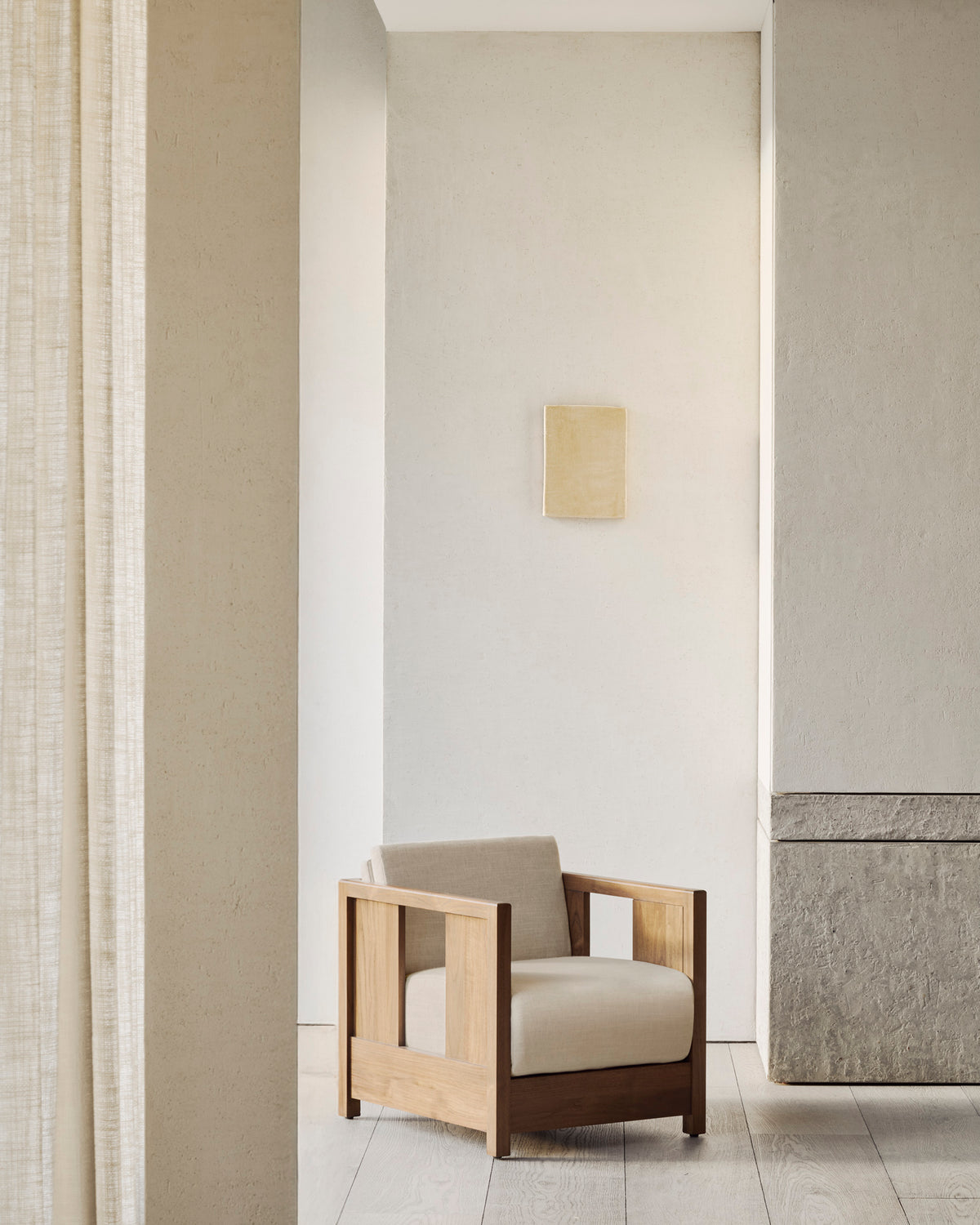 A minimalist room featuring the Ethnicraft Roan Lounge Chair in Flax, with beige cushions, light-colored walls, a stone panel, pale wood flooring, and a small square artwork on the wall above the chair.