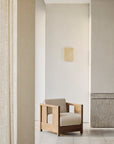 A minimalist room featuring the Ethnicraft Roan Lounge Chair in Flax, with beige cushions, light-colored walls, a stone panel, pale wood flooring, and a small square artwork on the wall above the chair.