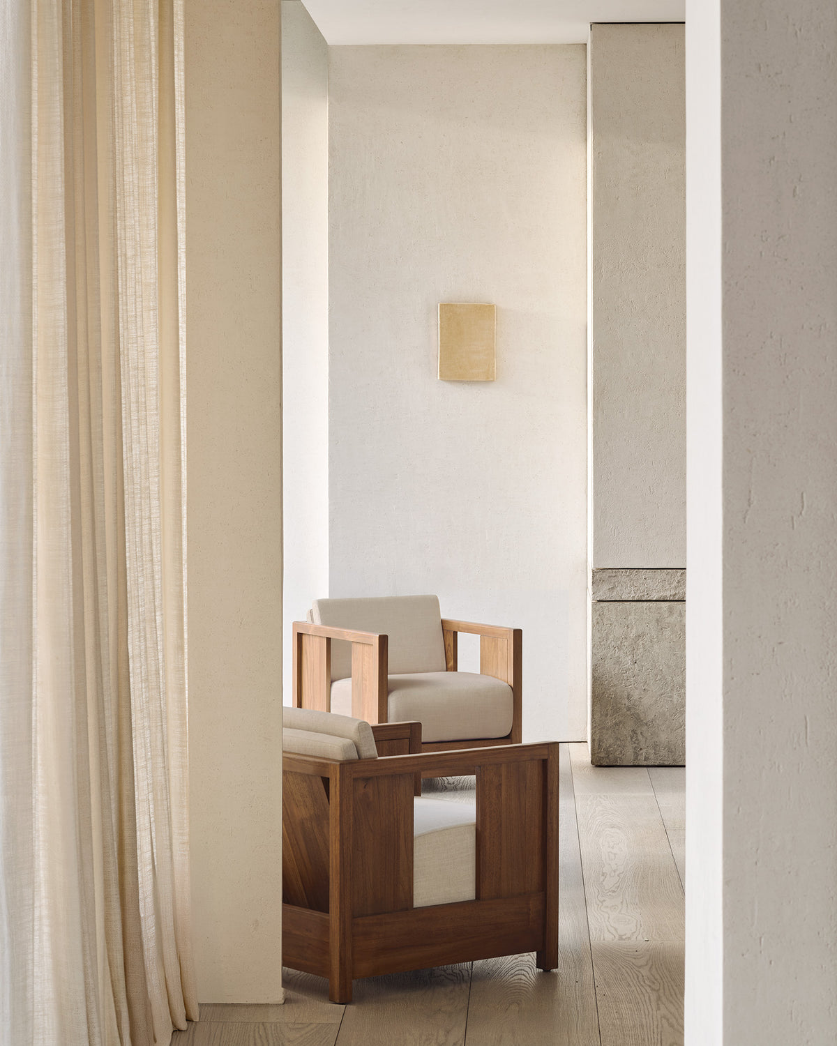 Minimalist room with light wood flooring, beige textured walls, and sheer curtains. A solid teak Roan Lounge Chair | Flax by Ethnicraft, adorned with light cushions, serves as the centerpiece. In the background, a small square wall art piece hangs. Soft natural lighting fills the space.