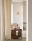 Minimalist room with light wood flooring, beige textured walls, and sheer curtains. A solid teak Roan Lounge Chair | Flax by Ethnicraft, adorned with light cushions, serves as the centerpiece. In the background, a small square wall art piece hangs. Soft natural lighting fills the space.