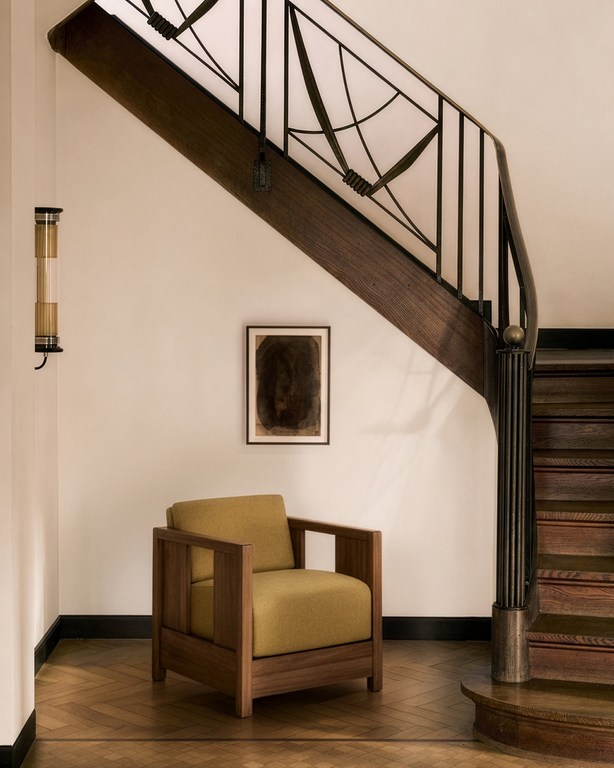 An Ethnicraft Roan Lounge Chair in Amber is placed beneath a staircase with a black railing. Above the chair, a framed abstract artwork adorns the wall, while an elegant wood herringbone pattern graces the floor below.