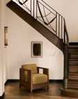 An Ethnicraft Roan Lounge Chair in Amber is placed beneath a staircase with a black railing. Above the chair, a framed abstract artwork adorns the wall, while an elegant wood herringbone pattern graces the floor below.