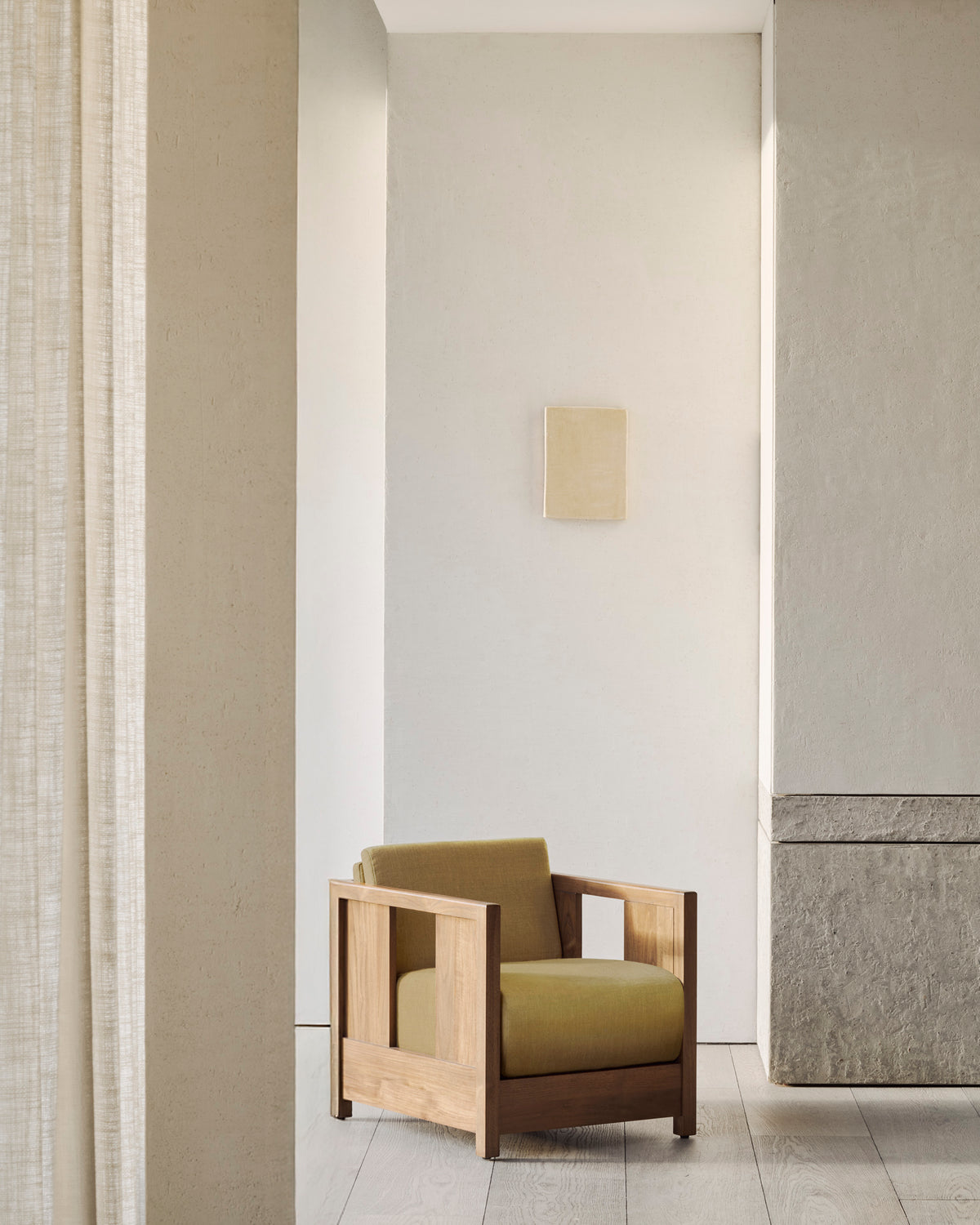 A modern room with light walls features the Roan Lounge Chair in Amber by Ethnicraft, adorned with olive green cushions and positioned beside a stone wall beneath a small, square wall art piece. Light filters in from the left, accentuating the minimalist decor.