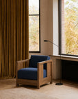 A modern Ethnicraft Roan Lounge Chair | Ink featuring deep navy cushions sits by large windows, golden curtains, and a black floor lamp in a cozy, minimalist room with light wood floors and autumn foliage visible outside.