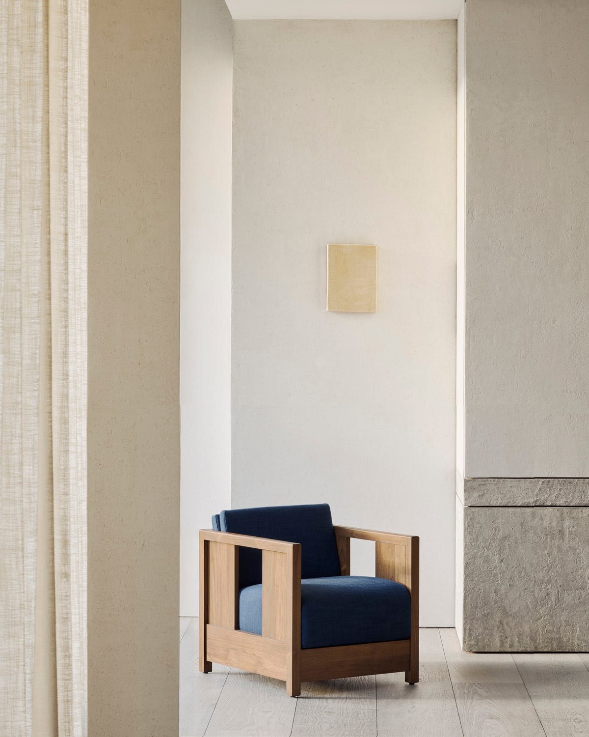The Roan Lounge Chair | Ink by Ethnicraft, with its vintage-inspired modern wooden frame and blue cushions, is featured in a minimalist room with light-colored walls, a small square artwork, beige curtains, and textured stone accents.