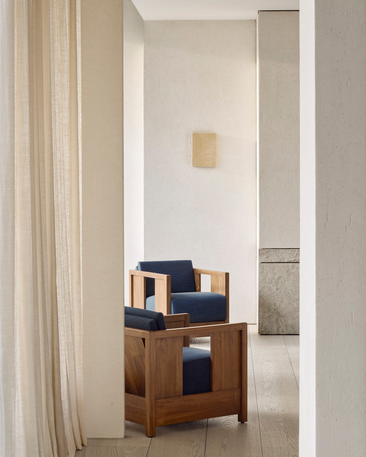 Two Ethnicraft Roan Lounge Chairs in Ink are placed in a minimalist, light-filled room with beige walls, sheer curtains, and a wall-mounted light fixture.