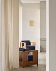 Two Ethnicraft Roan Lounge Chairs in Ink are placed in a minimalist, light-filled room with beige walls, sheer curtains, and a wall-mounted light fixture.