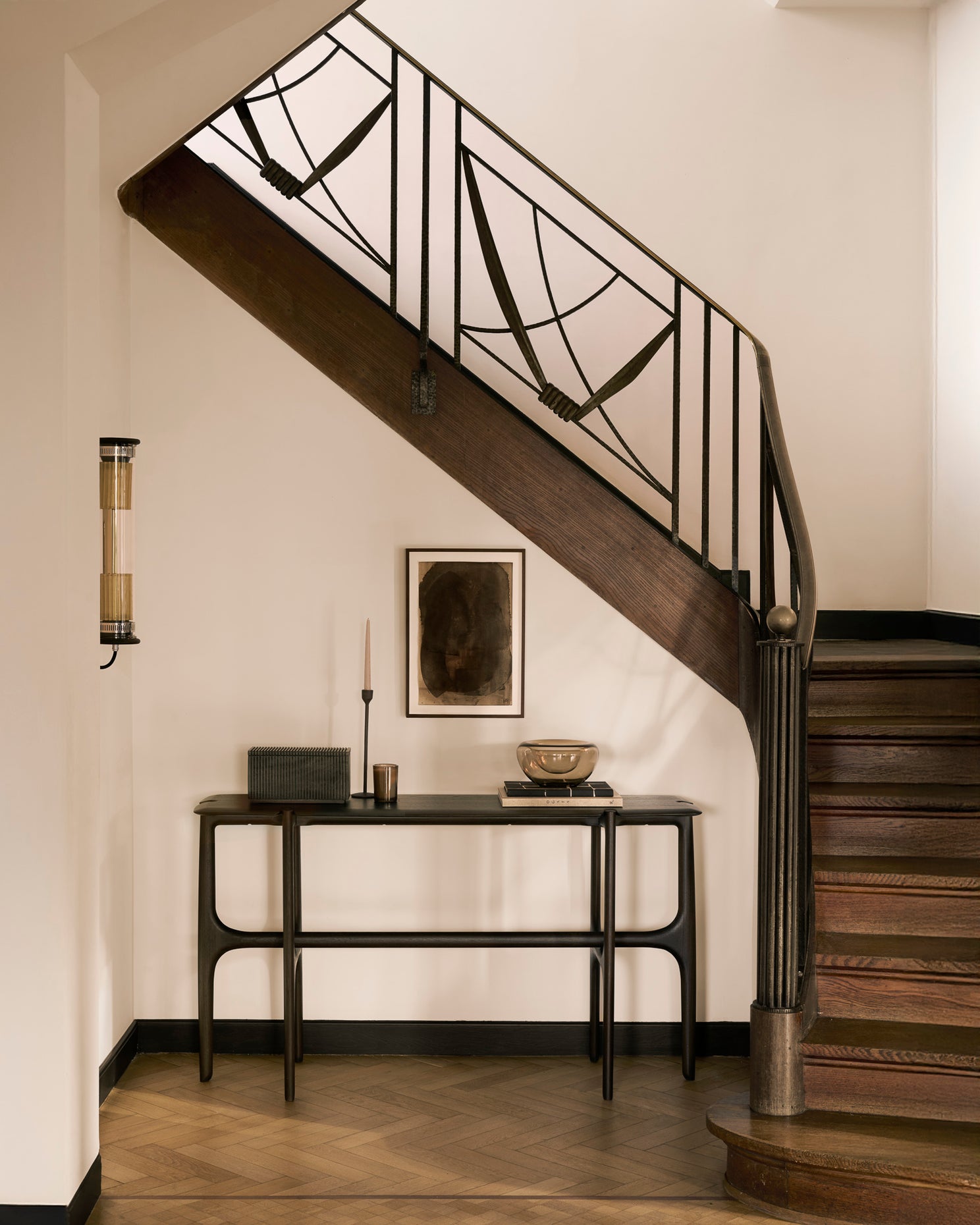 A wooden staircase with black metal railings curves above a minimalist Ethnicraft PI Console in dark brown teak. Decorative accents and abstract framed art enhance the space’s neutral palette.
