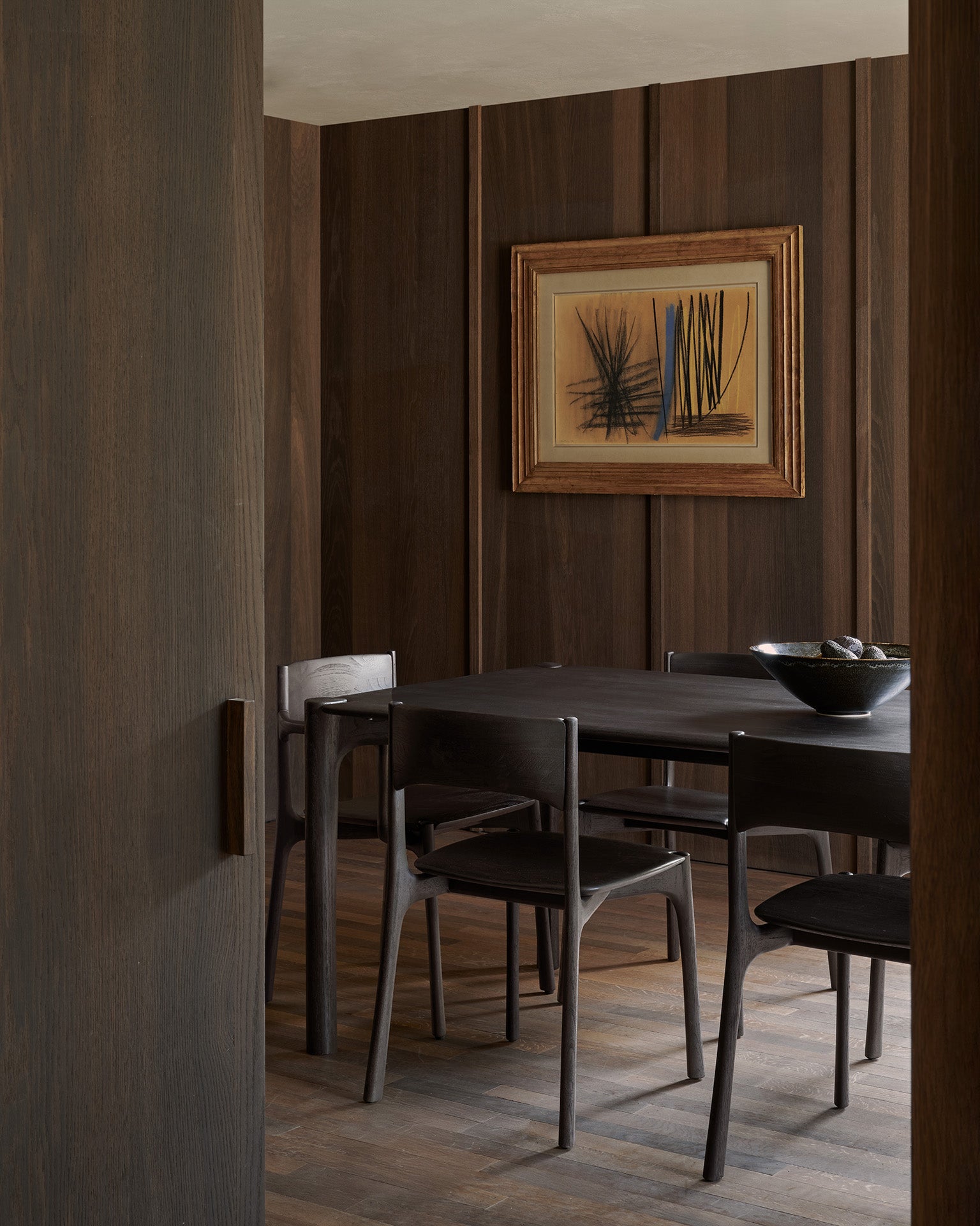 A minimalist dining room with dark wood walls, a black rectangular table surrounded by five Ethnicraft PI Dining Chairs in Teak Dark Brown, a bowl centerpiece, and abstract framed artwork on the wall.