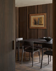 A modern dining room with dark wood paneling features a black dining table surrounded by four Ethnicraft PI Dining Chairs in Teak Dark Brown with arms, a bowl centerpiece, and abstract framed artwork. The scene is viewed through an open doorway.