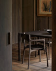 A dimly lit room with wood-paneled walls features a dark wooden dining table and the Ethnicraft PI Dining Chair in Teak Dark Brown with arms. A contemporary framed artwork is displayed above the table.
