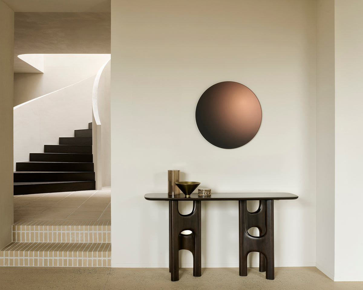 An Ethnicraft Flow Console | Mahogany Rose Brown with cut-out legs stands against a wall beneath a large circular mirror with a gradient finish.