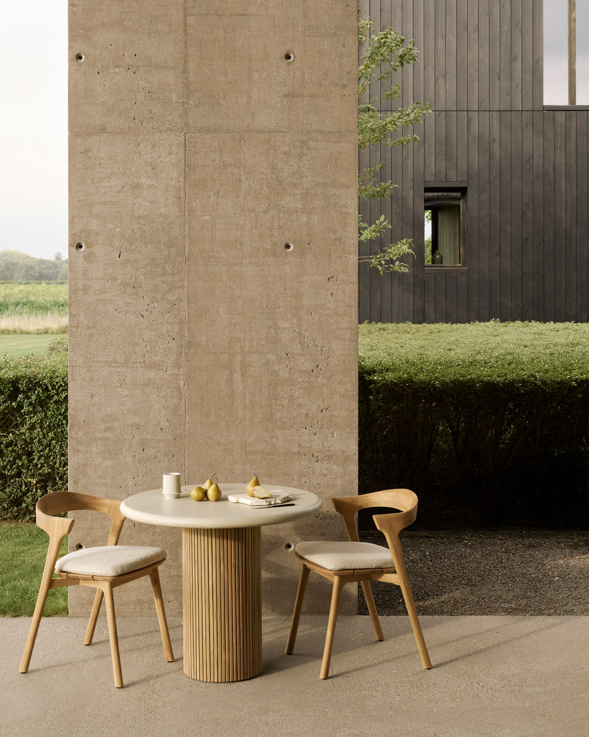 A small, round Roller Max Outdoor Dining Table by Ethnicraft with a hand-finished concrete top is set outdoors beside a concrete wall, accompanied by two wooden chairs. Fruit and tableware are arranged on the table, while bushes and a modern dark building serve as the backdrop.