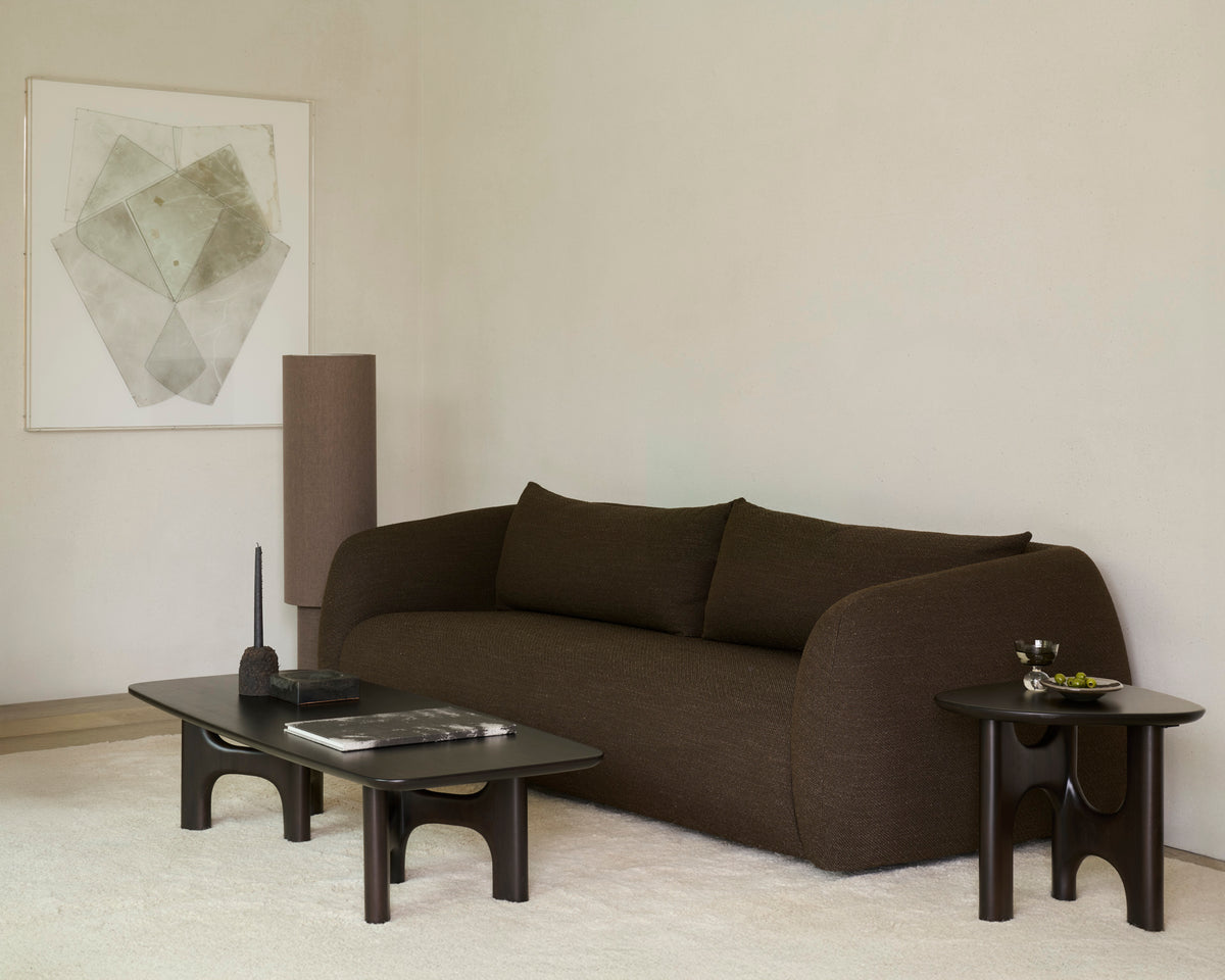 Minimalist living room with a dark brown sofa, two vintage modern furniture pieces—including an Ethnicraft Flow Side Table and a solid mahogany side table—a tall cylindrical lamp, abstract wall art, and a light textured rug on neutral-colored floor and walls.