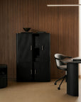 Modern office with wood-paneled walls, an Ethnicraft Roller Max Filing Cabinet in black, a matching black desk, a swivel chair, and minimal décor, including a black bowl and a sleek linear light for stylish and functional storage.
