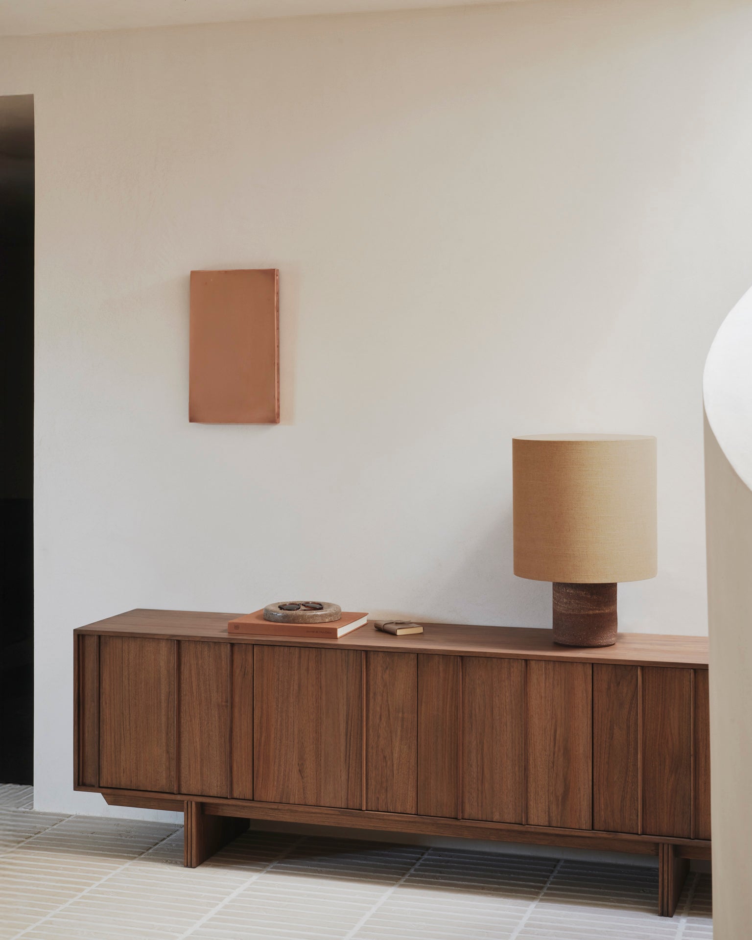 A minimalist room featuring Ethnicraft’s Pillar Sideboard in Teak Wild Brown, a beige table lamp, a book, and decor objects. A small rectangular artwork hangs above the sideboard on a light wall; the floor displays a subtle tiled pattern.