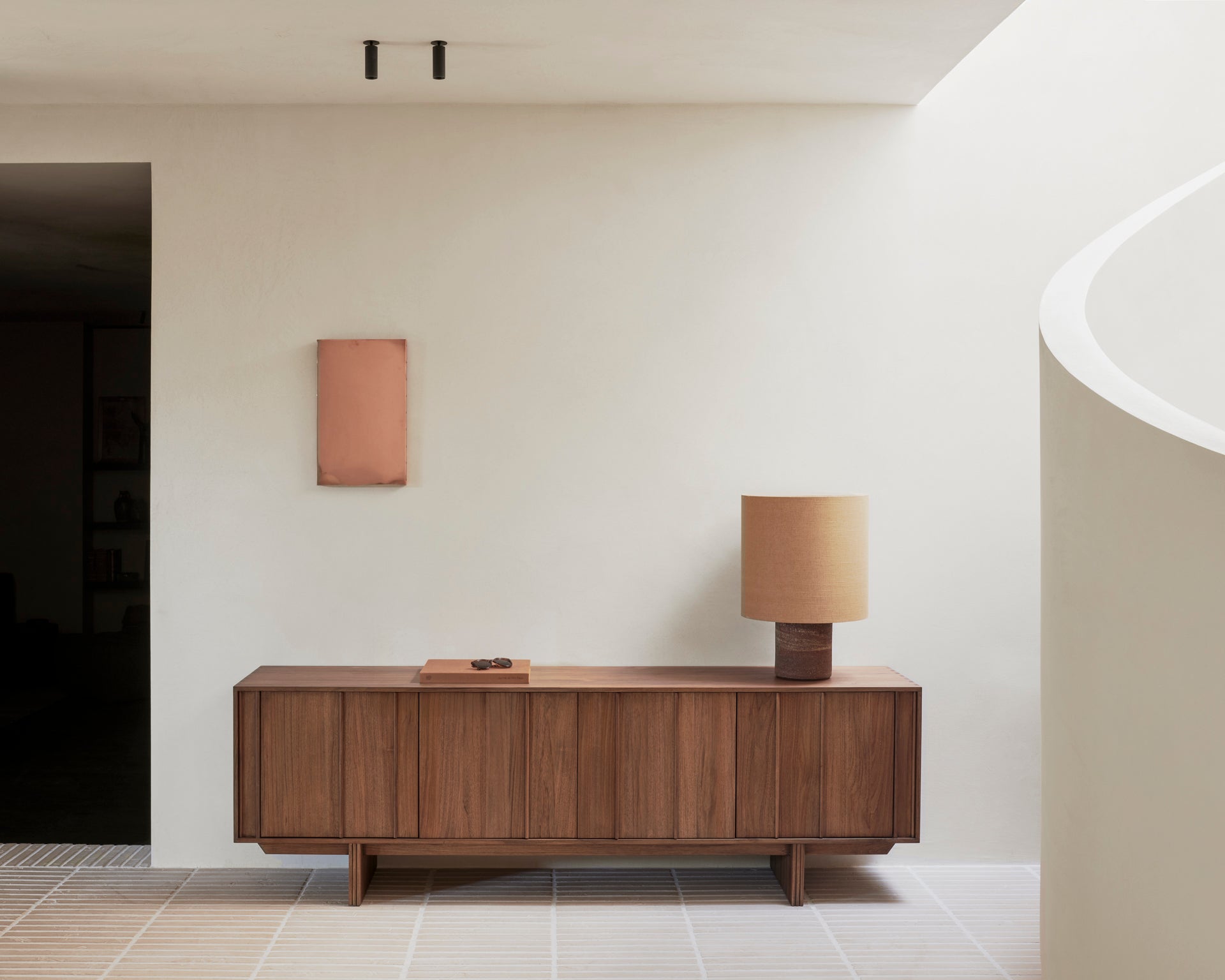 A minimalist interior with the Ethnicraft Pillar Sideboard - Teak Wild Brown, a tan table lamp, and rectangular pink wall art on a white wall. Natural light from the right enhances the clean, modern style.