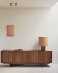 A minimalist interior with the Ethnicraft Pillar Sideboard - Teak Wild Brown, a tan table lamp, and rectangular pink wall art on a white wall. Natural light from the right enhances the clean, modern style.