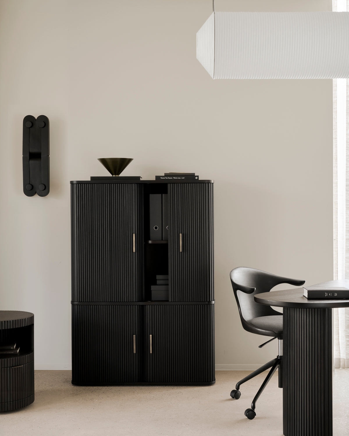 Modern office featuring black ribbed furniture, a swivel chair, round table, and the Ethnicraft Roller Max Filing Cabinet for stylish storage. Minimalist decor includes a geometric wall lamp, abstract bowl, and a hanging white rectangular light fixture. Neutral tones on the walls and floor complete the look.