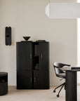 Modern office featuring black ribbed furniture, a swivel chair, round table, and the Ethnicraft Roller Max Filing Cabinet for stylish storage. Minimalist decor includes a geometric wall lamp, abstract bowl, and a hanging white rectangular light fixture. Neutral tones on the walls and floor complete the look.