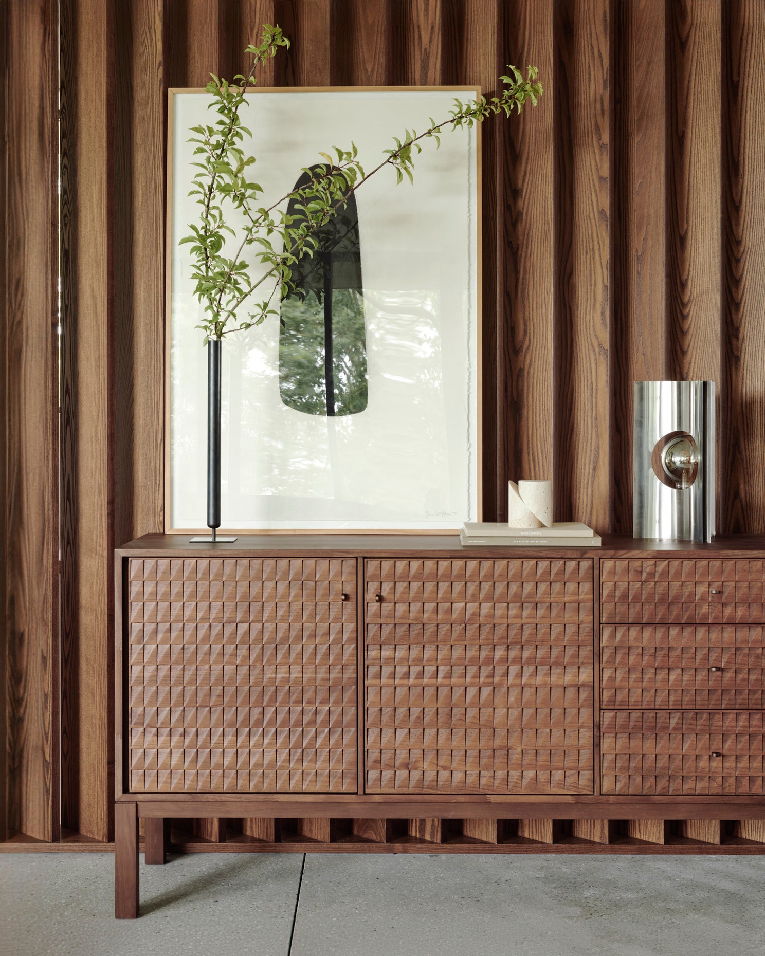 The Ethnicraft Sono Sideboard, with its woven front and classic European refinement, stands against a wood-paneled wall topped with a tall vase of green branches, framed abstract artwork, a small box, and a cylindrical metallic sculpture.