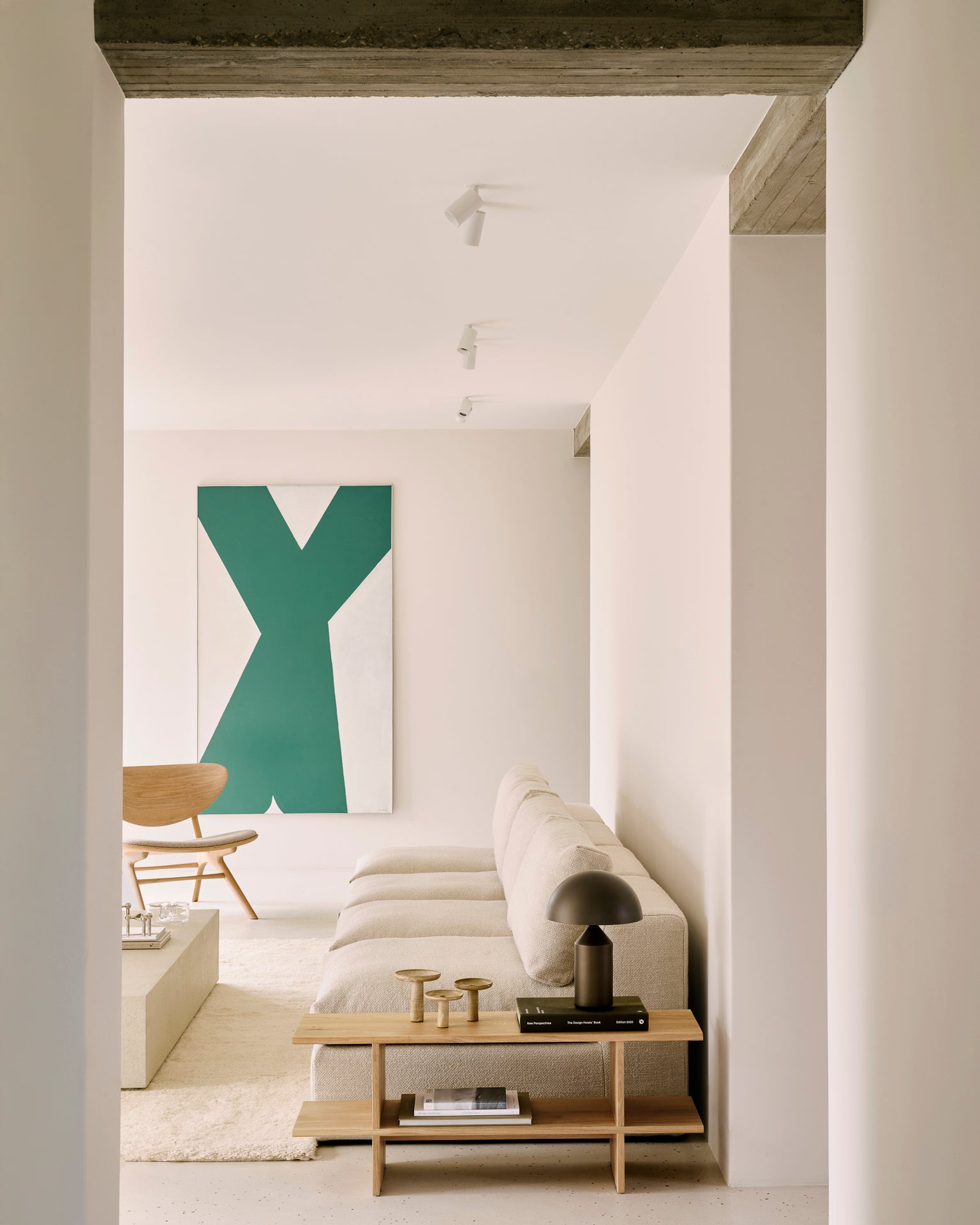 A minimal living room features a beige sectional sofa, wooden coffee table, black lamp, décor pieces, and a large green and white abstract painting above a wooden chair and the Ethnicraft Kabuki Side Table - Oak.
