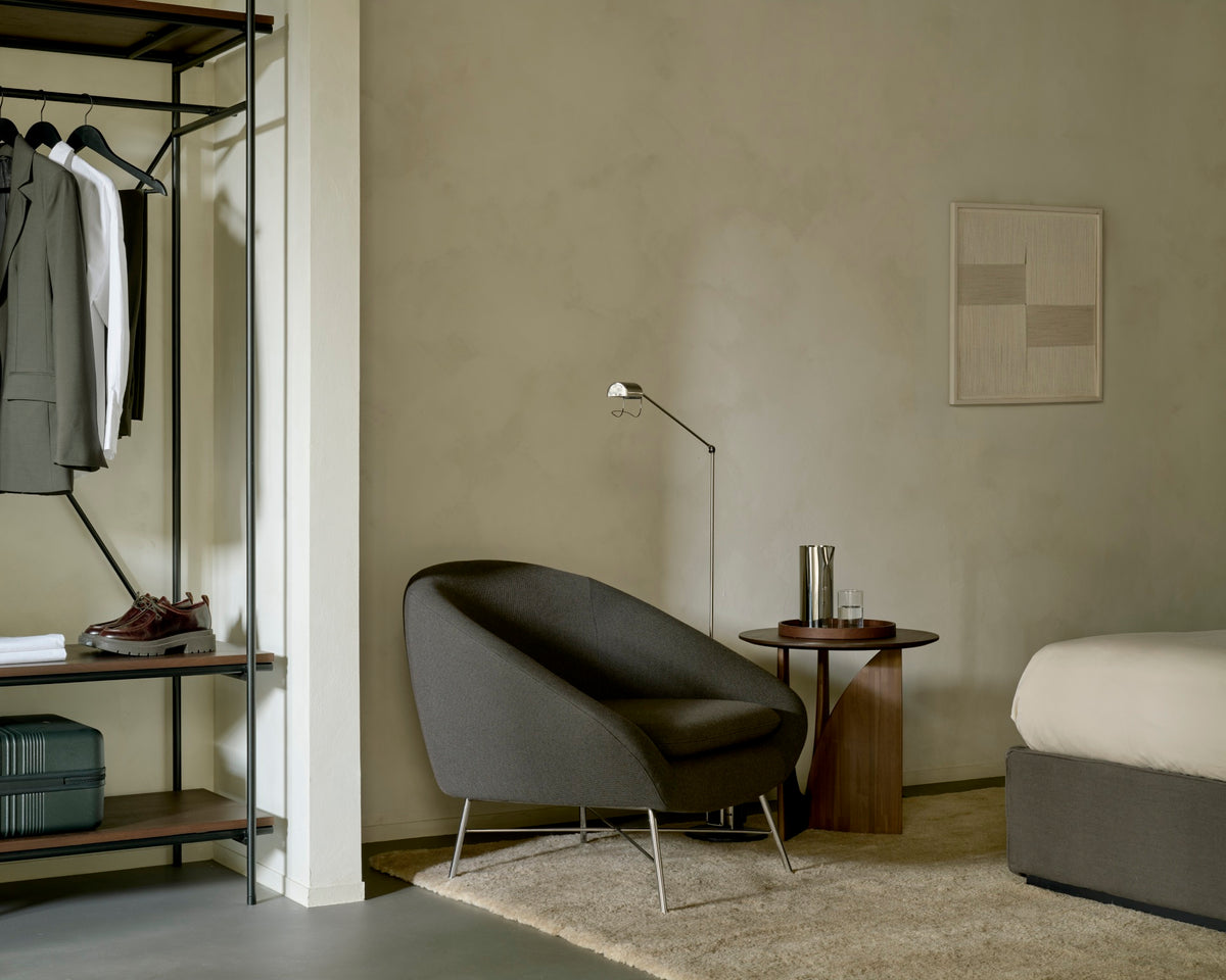 A dark gray Ethnicraft Barrow Lounge Chair - Paon with a curved back and thin metal legs sits next to a small wooden side table in a room with a metal clothing rack.