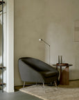 A dark gray Ethnicraft Barrow Lounge Chair - Paon with a curved back and thin metal legs sits next to a small wooden side table in a room with a metal clothing rack.
