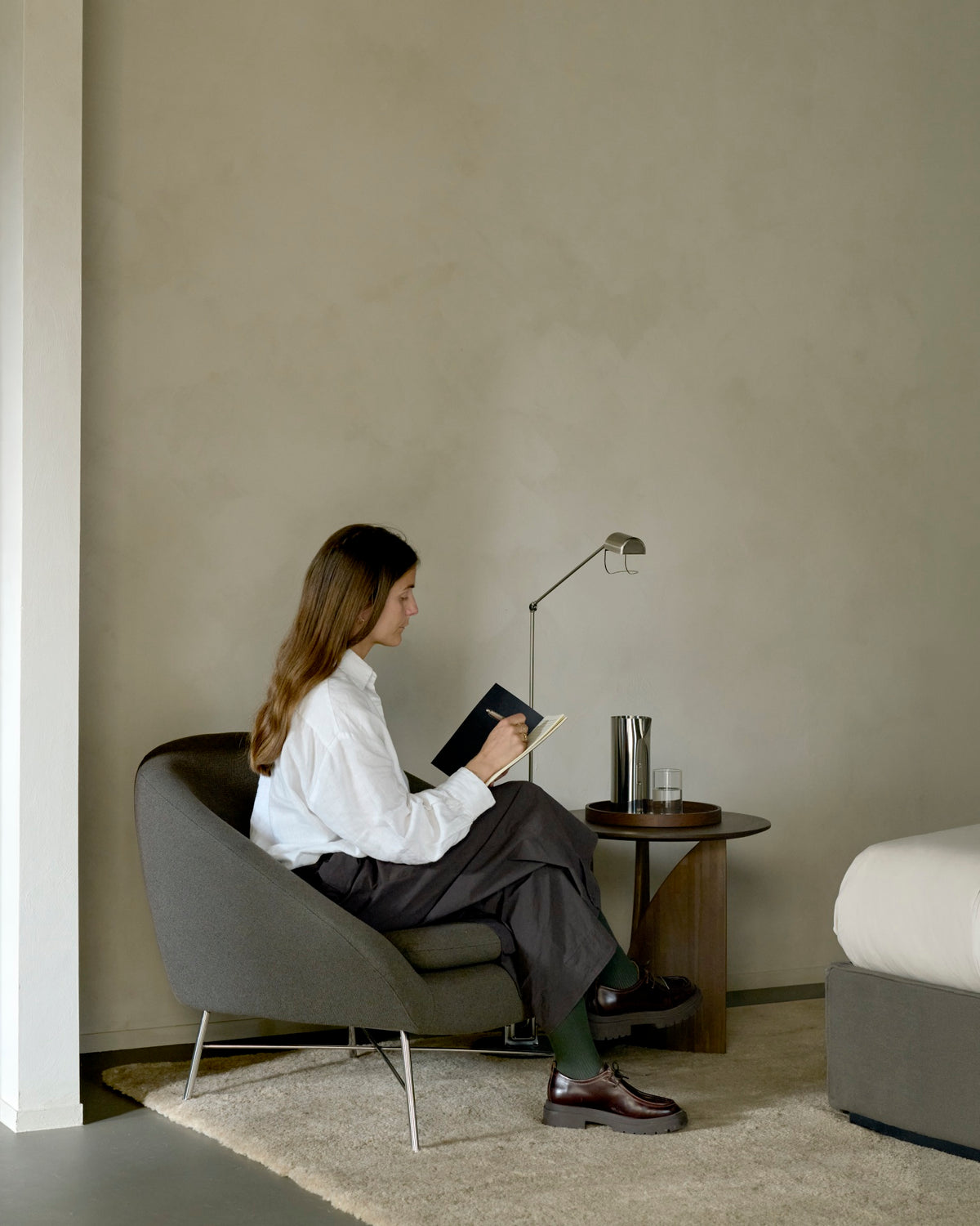 A person sits in a gray Ethnicraft Barrow Lounge Chair - Paon, writing in a notebook next to a small side table.