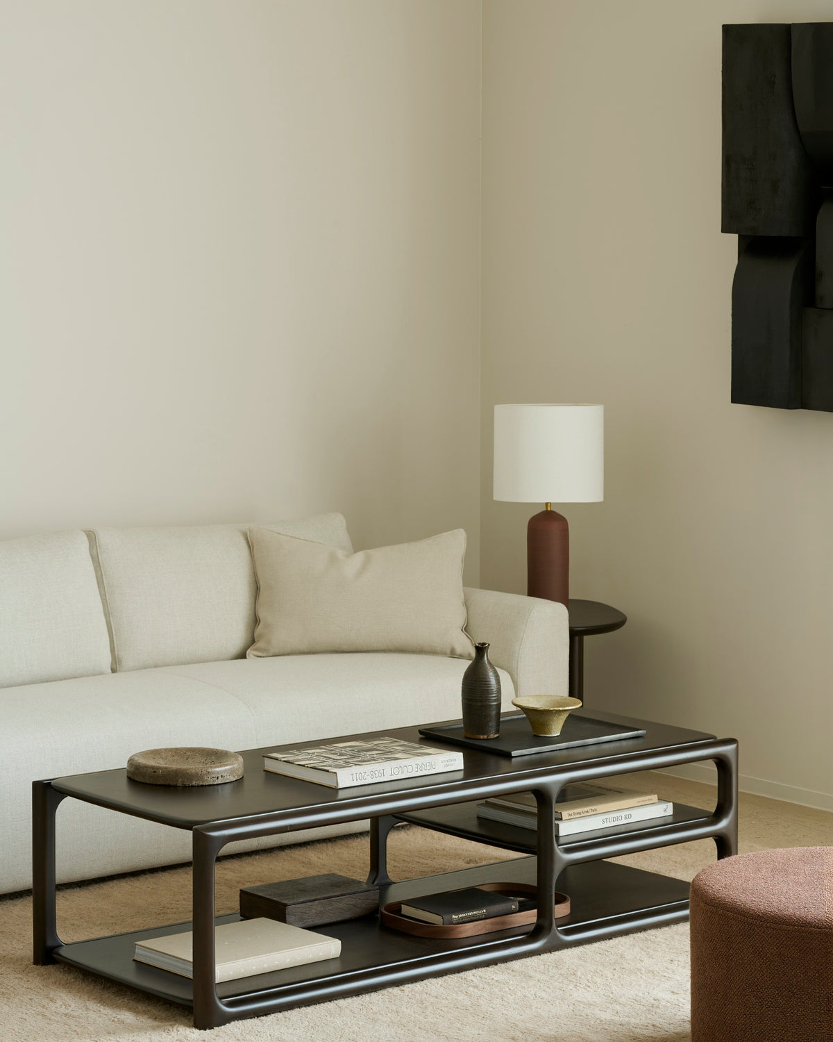 A minimalist living room featuring a light beige sofa, a neutral pillow, the Ethnicraft Eda Coffee Table adorned with books and decor, a round mahogany occasional table with a brown vase lamp, and abstract wall art—all showcasing heirloom-quality furniture throughout the space.