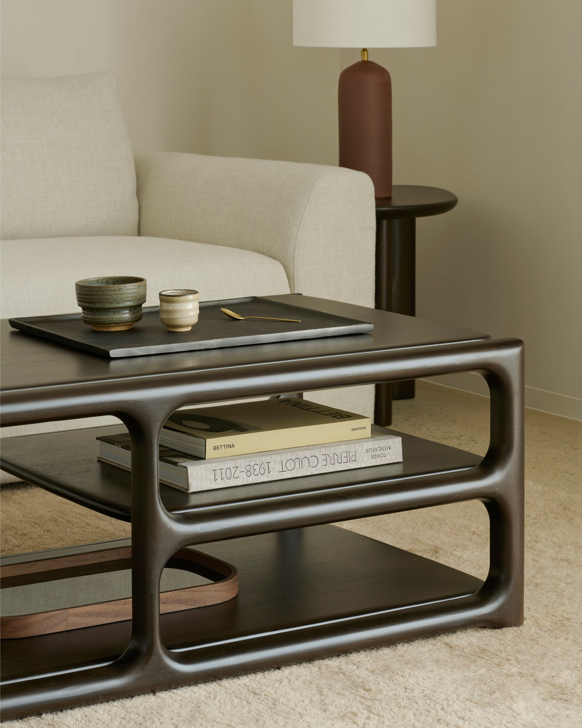 A minimalist living room features a cream sofa and an Ethnicraft Eda Coffee Table in dark wood, adorned with ceramic cups, a book, and a tray. Beside the sofa stands an Ethnicraft mahogany occasional table with a brown lamp, all arranged atop a beige carpet.
