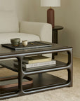 A minimalist living room features a cream sofa and an Ethnicraft Eda Coffee Table in dark wood, adorned with ceramic cups, a book, and a tray. Beside the sofa stands an Ethnicraft mahogany occasional table with a brown lamp, all arranged atop a beige carpet.