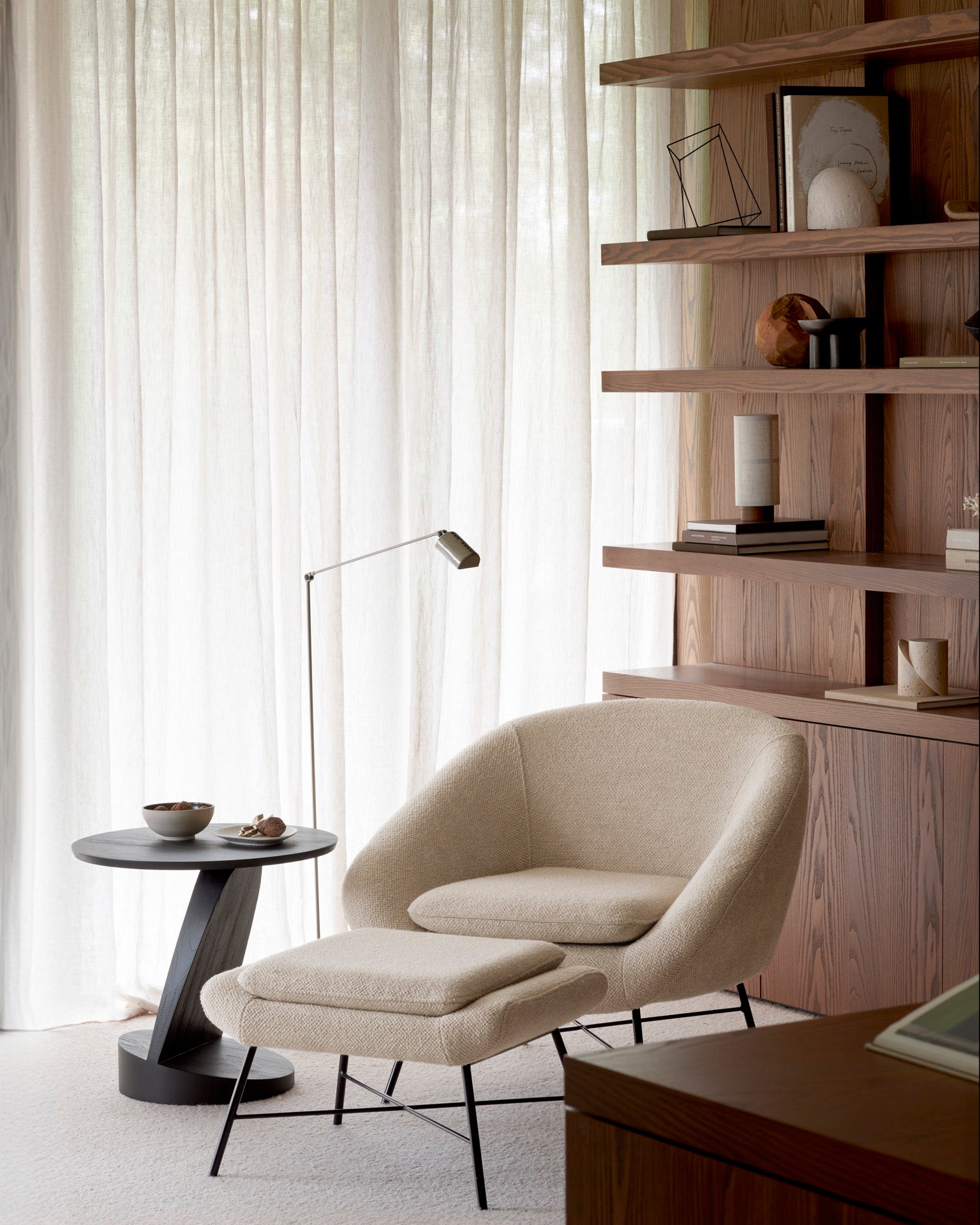 A cozy reading nook featuring the Barrow Lounge Chair - Off White by Ethnicraft, paired with a matching ottoman, a round side table, floor lamp, and wooden shelves filled with books and decor accents—all set against sheer white curtains and a warm wood-paneled wall.