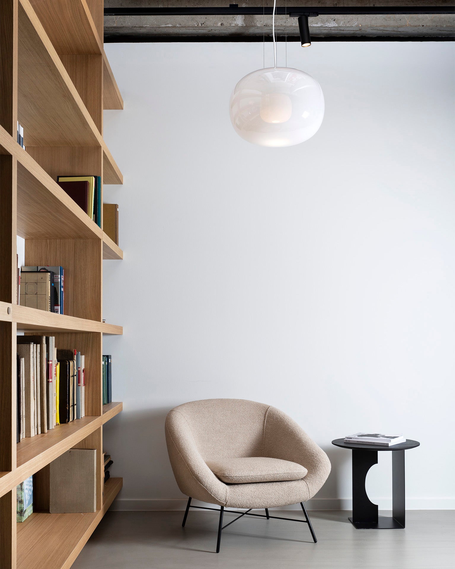 A modern reading nook featuring the Ethnicraft Barrow Lounge Chair in off white, a petite black side table with stacked books, wooden Ethnicraft shelves filled with books, and a white pendant light suspended from the ceiling against a crisp white wall.
