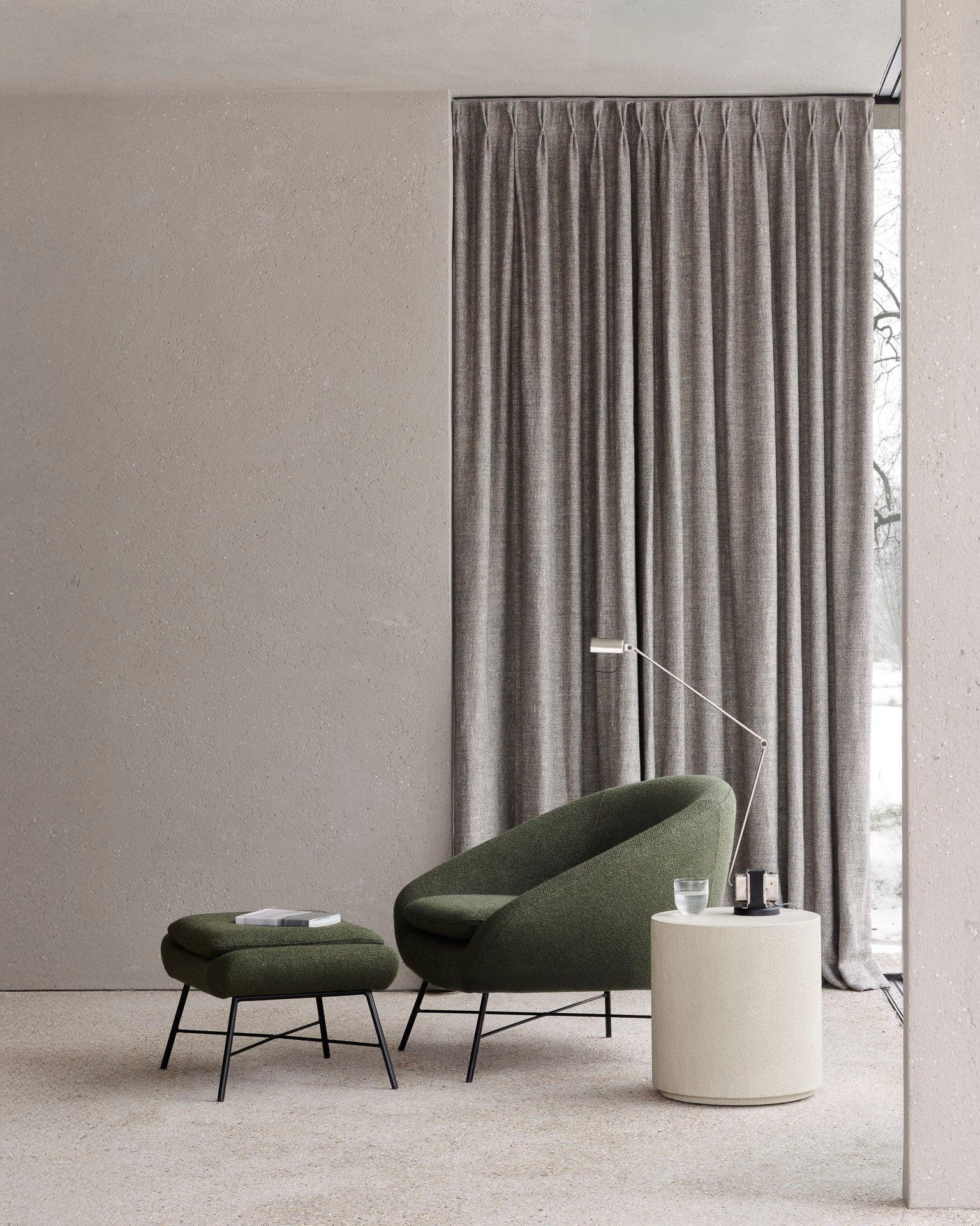 A modern living space with a pine green Barrow Footstool by Ethnicraft, complemented by a lounge chair, round side table, floor lamp, and tall gray curtains. A book and glass of water add to the minimalist, stylish setting.