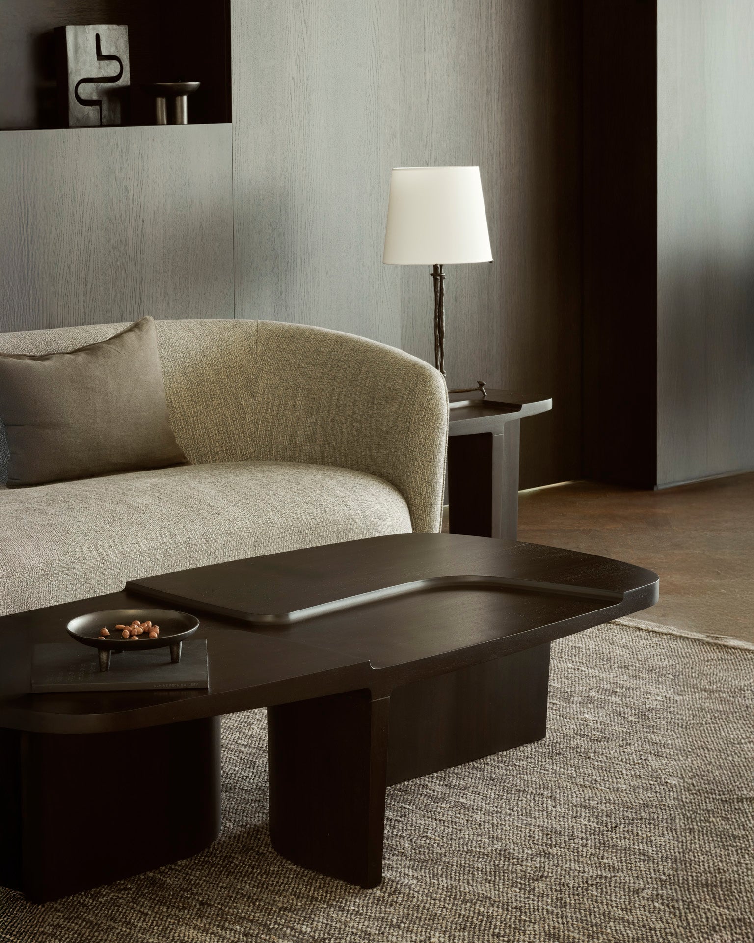 A modern living room features a curved gray sofa, an Ethnicraft Brutalist Coffee Table holding a small dish of nuts and tray, a side table with a white lamp, plus neutral-toned walls and carpet.