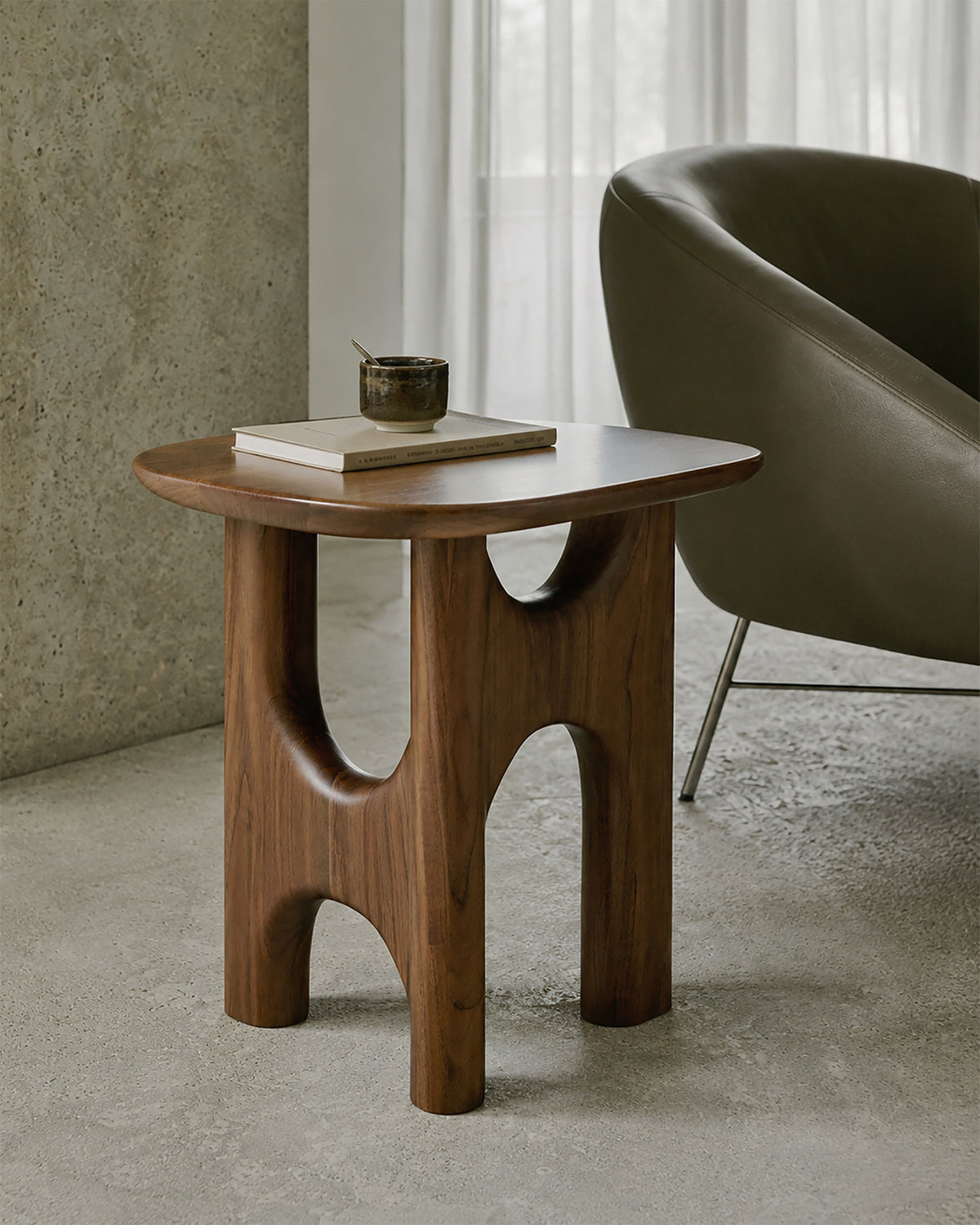 The Flow Side Table by Ethnicraft—a modern piece crafted from solid mahogany with sculptural legs—holds a book and a cup as it stands on a concrete floor beside a gray armchair and a window with sheer curtains, making a refined statement of vintage modern furniture.