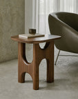 The Flow Side Table by Ethnicraft—a modern piece crafted from solid mahogany with sculptural legs—holds a book and a cup as it stands on a concrete floor beside a gray armchair and a window with sheer curtains, making a refined statement of vintage modern furniture.