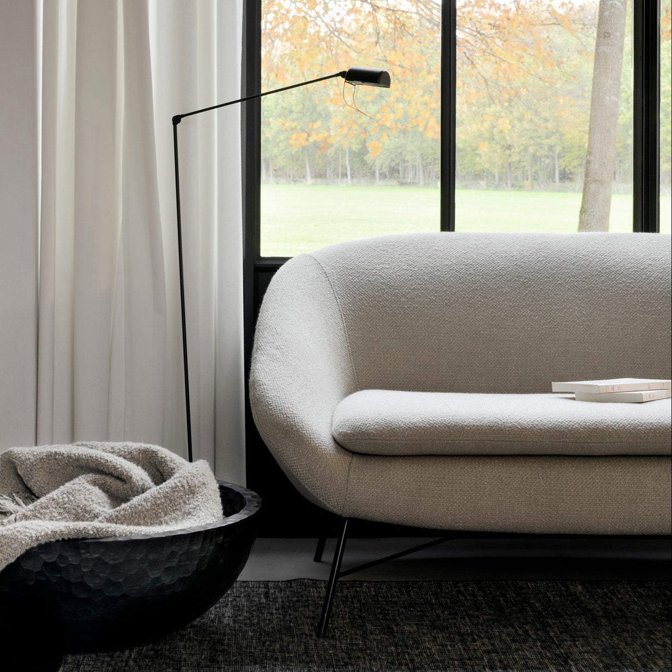 A modern living room features a curved light beige sofa, a black floor lamp, and a black textured bowl with a gray throw beside large windows, accented by Ethnicrafts Dots Kilim Rug that adds warmth to the autumn view outside.