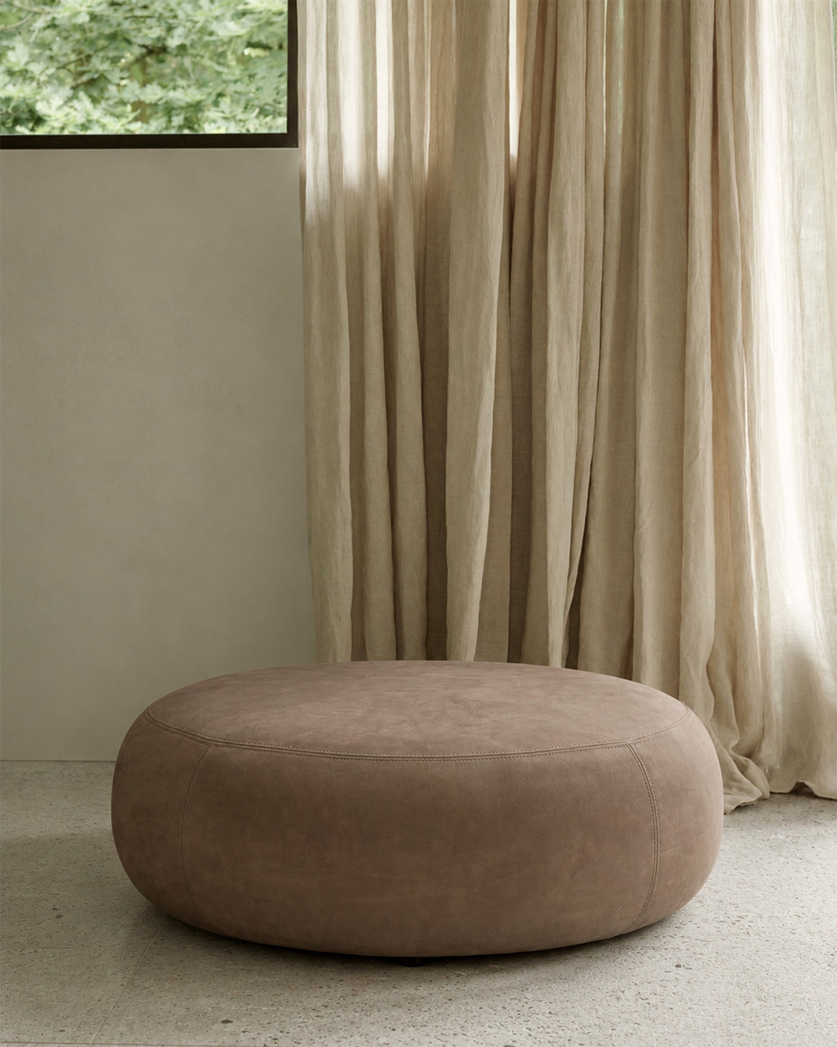A low, round Ethnicraft Orb Pouf | Fawn Nubuck Leather pouf in brown aniline leather upholstery sits on a stone floor.