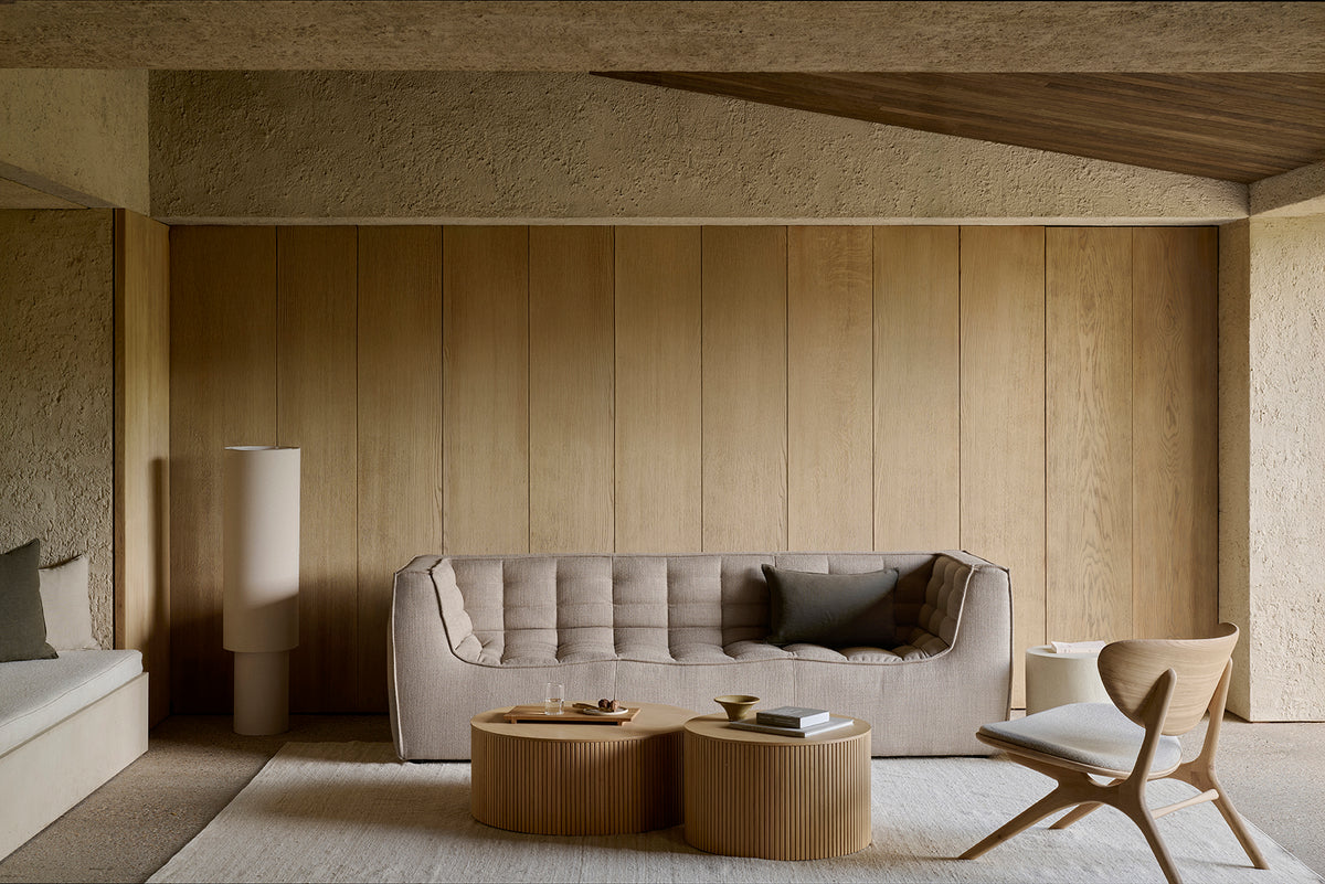 Minimalist living room with neutral tones, featuring the Ethnicraft N701 3-Seater Sofa in beige tufted upholstery, round wooden coffee tables, a sculptural wooden chair, floor lamp, and textured wood panel walls. A light rug covers the floor, highlighting timeless European design influences.