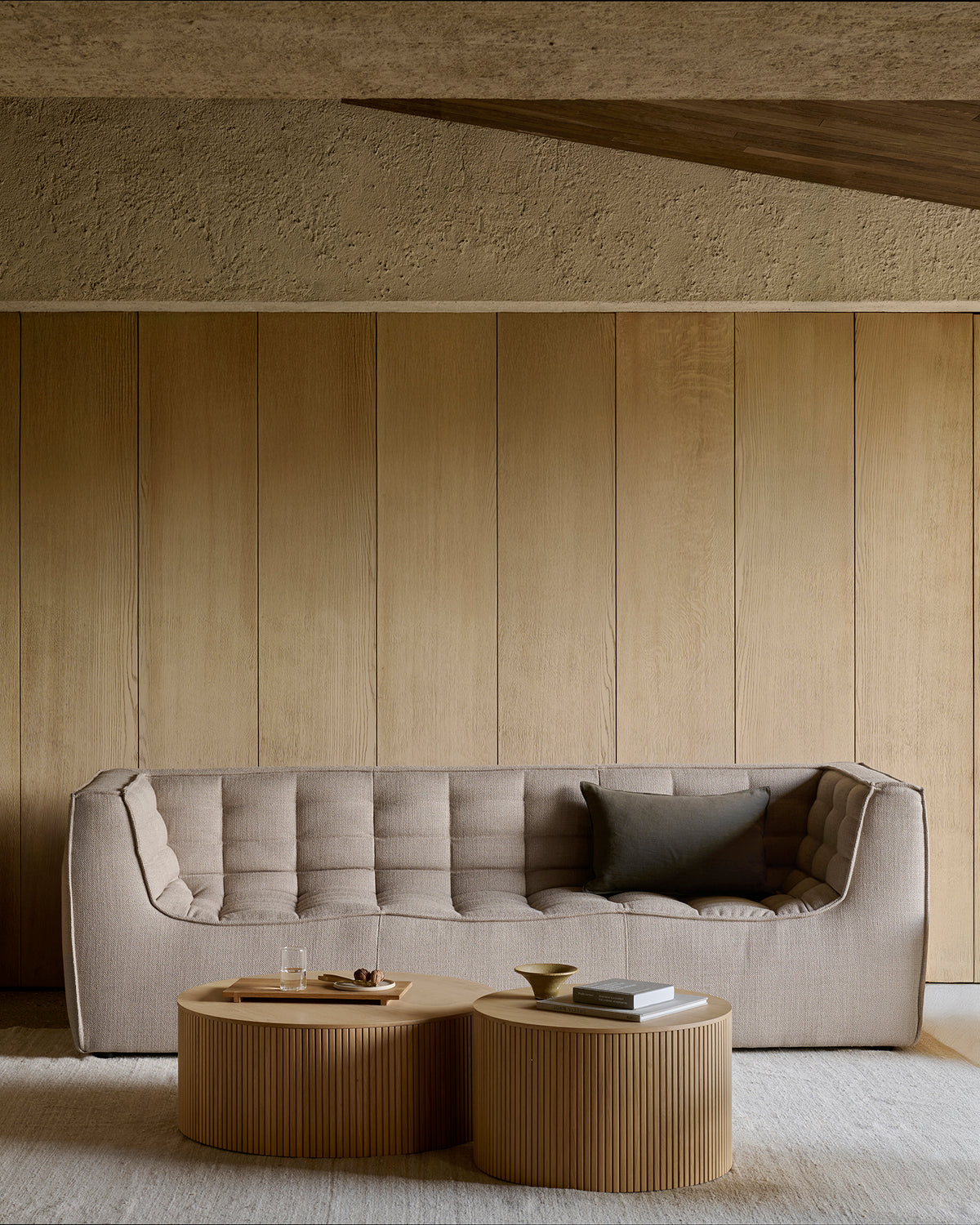 An N701 3-Seater Sofa in beige by Ethnicraft, accented with a black pillow, sits against a wooden panel wall, showcasing elegant European design. In front, two round wooden coffee tables display a tray, glasses, a bowl, and books. The space features a minimalist aesthetic with neutral tones.