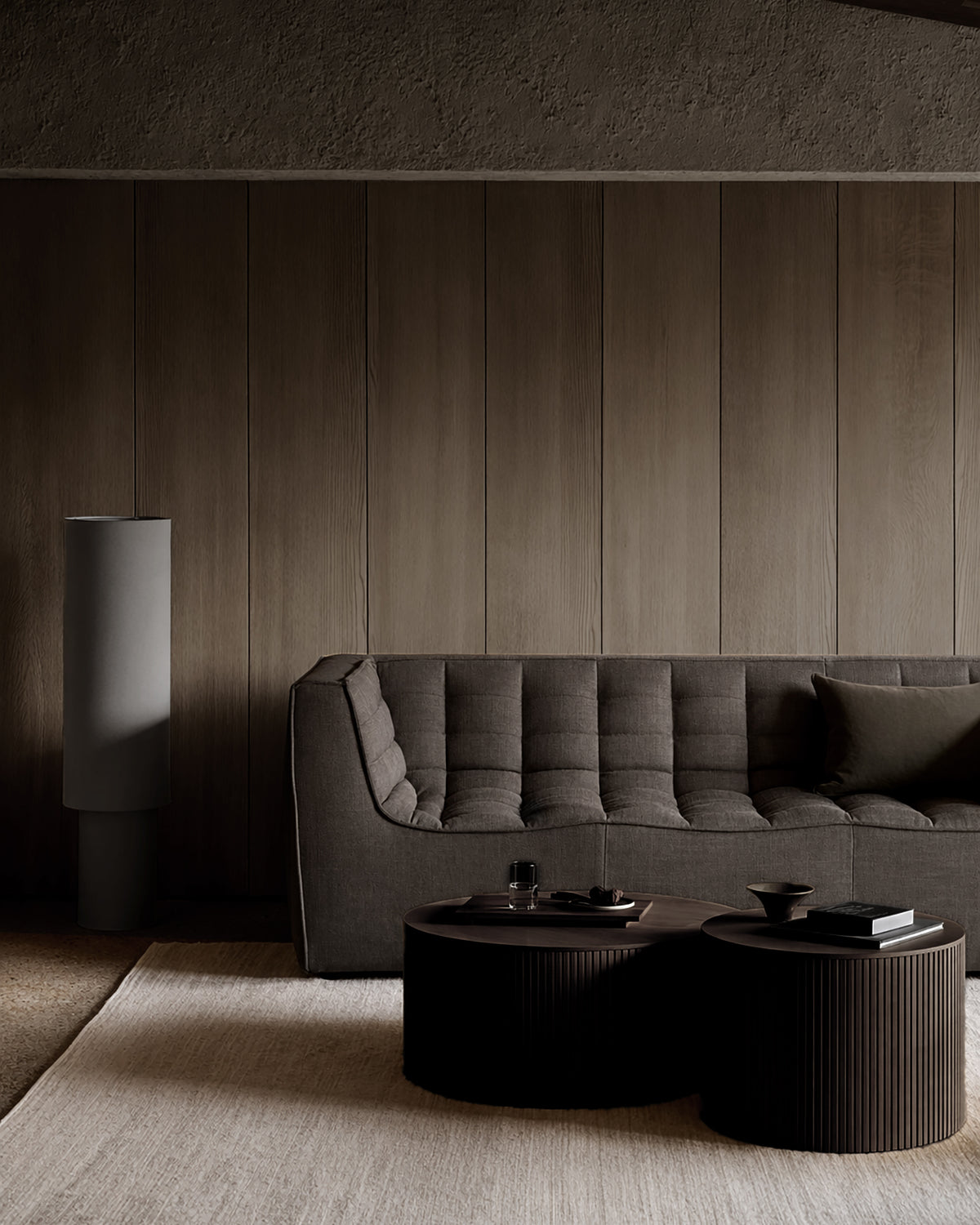 Minimalist living room featuring the Ethnicraft N701 3-Seater Sofa in Dark Grey with a matching pillow, two round coffee tables styled with books and cups, a textured beige rug, and a tall cylindrical lamp set against a paneled wooden wall.
