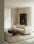 A modern minimalist living room showcases the Ethnicraft Figur Sofa in white, a beige armchair, black coffee table, books, and a tall dark shelf on a light rug with sheer curtains—ideal for contemporary living.