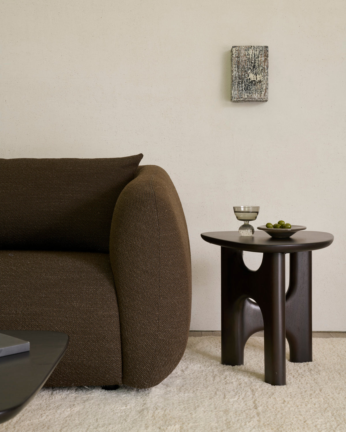 An Ethnicraft Flow Side Table | Mahogany with a sculptural base, holding a small bowl of olives and a glass of water next to a brown sofa.