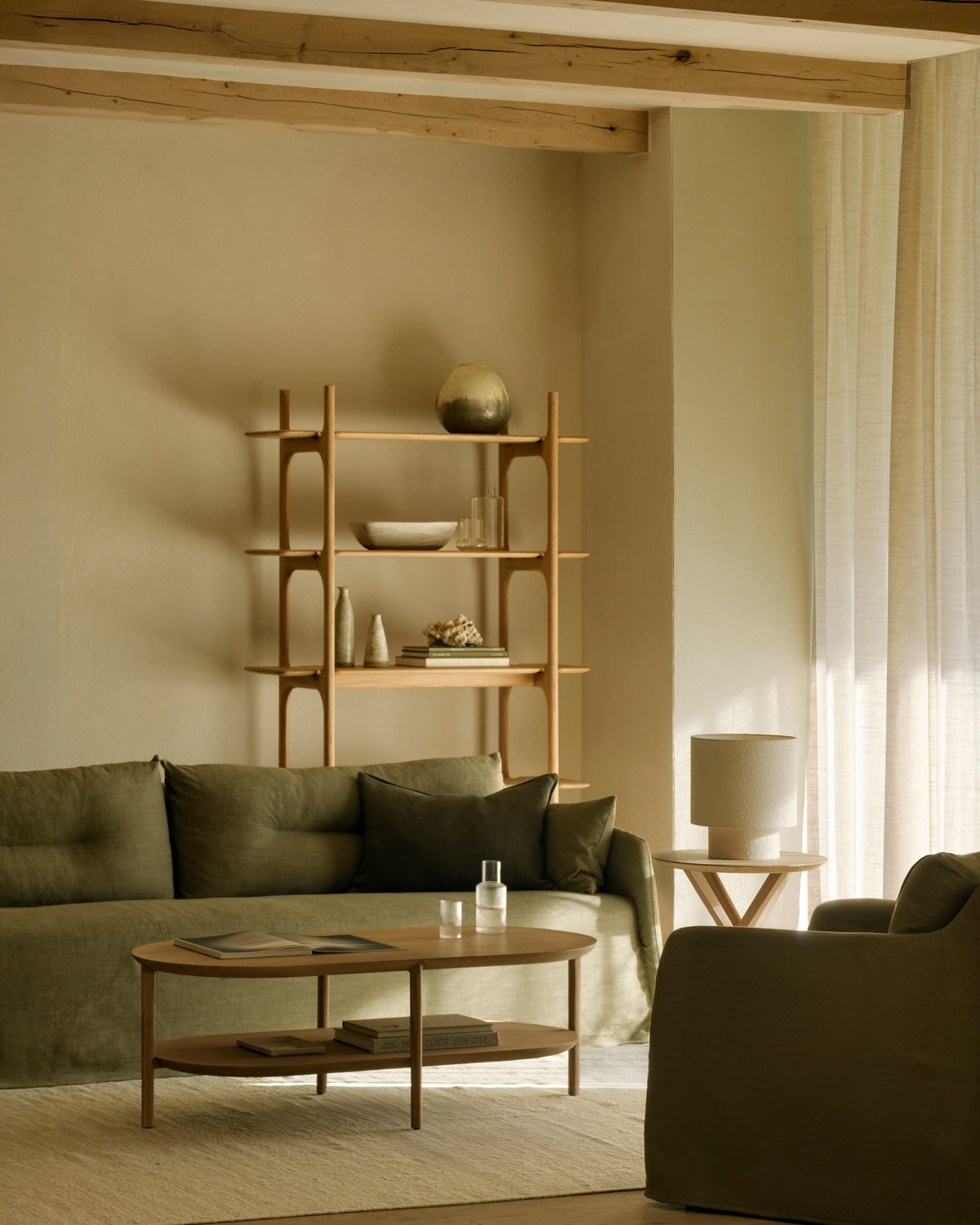 A minimalist living room featuring the Ethnicraft Weave Sofa | 2 Seater Green with its olive green, linen-blend upholstery stands alongside a wooden coffee table and a simple wooden shelf decorated with curated items. A side table with a lamp, light curtains, and exposed ceiling beams complete the look, while neutral tones create a calm and cozy atmosphere.