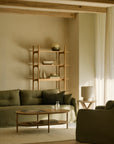 A minimalist living room featuring the Ethnicraft Weave Sofa | 2 Seater Green with its olive green, linen-blend upholstery stands alongside a wooden coffee table and a simple wooden shelf decorated with curated items. A side table with a lamp, light curtains, and exposed ceiling beams complete the look, while neutral tones create a calm and cozy atmosphere.